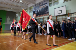 Na zdjęciu uczniowie ubrani na galowo na sali gimnastycznej, dwóch chłopców niesie czerwony sztandar szkoły im. A. Mickiewicza