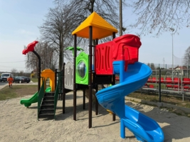 Na zdjęciu duża zabawka dla dzieci, ze schodkami, zjezdżalniami, daszkiem i przejściem, z tyłu ścianka wspinaczkowa